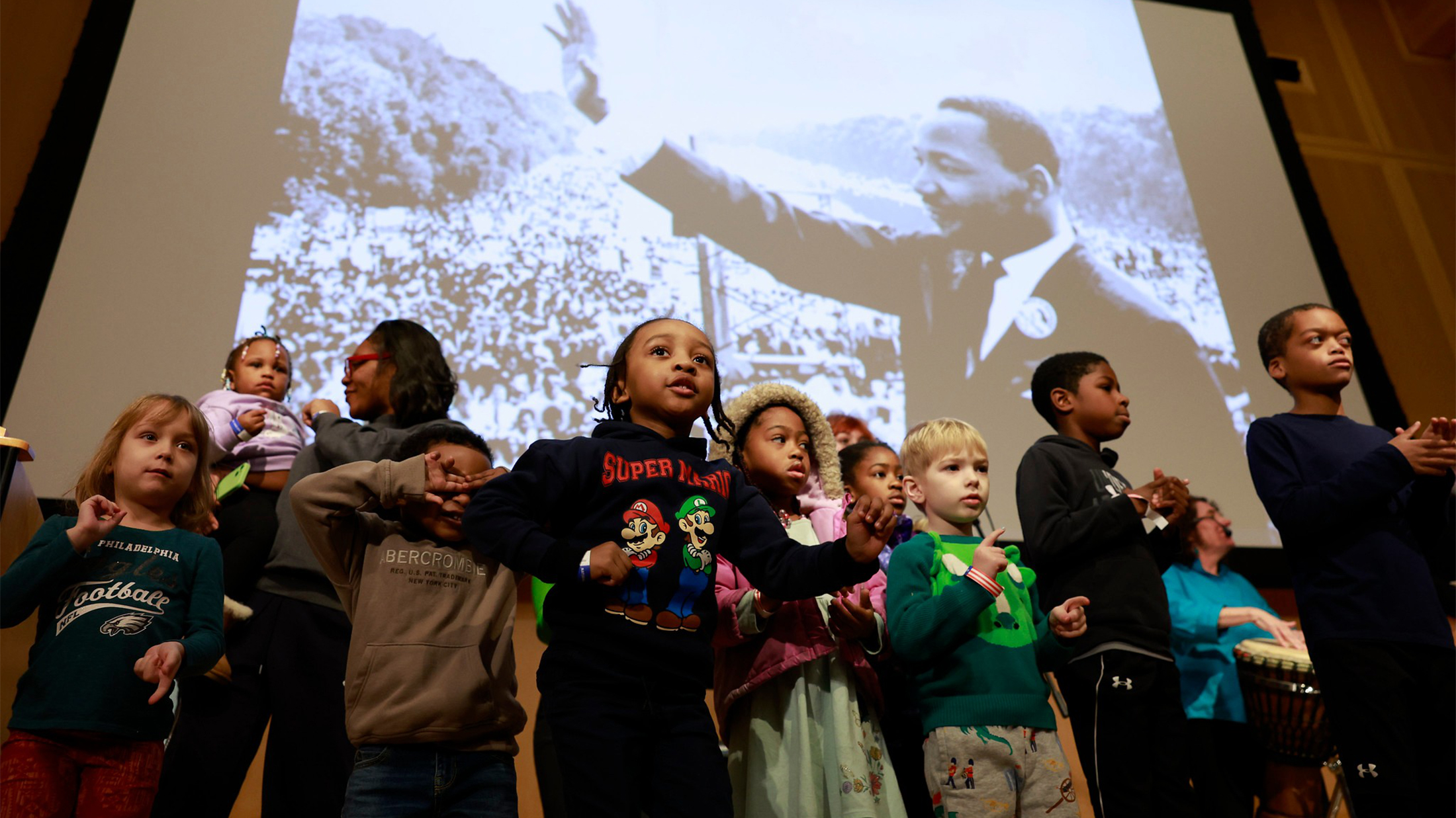 A group of children stands onstage in front of a large projected photo of Martin Luther King Jr., with several kids clapping and moving as part of a performance.