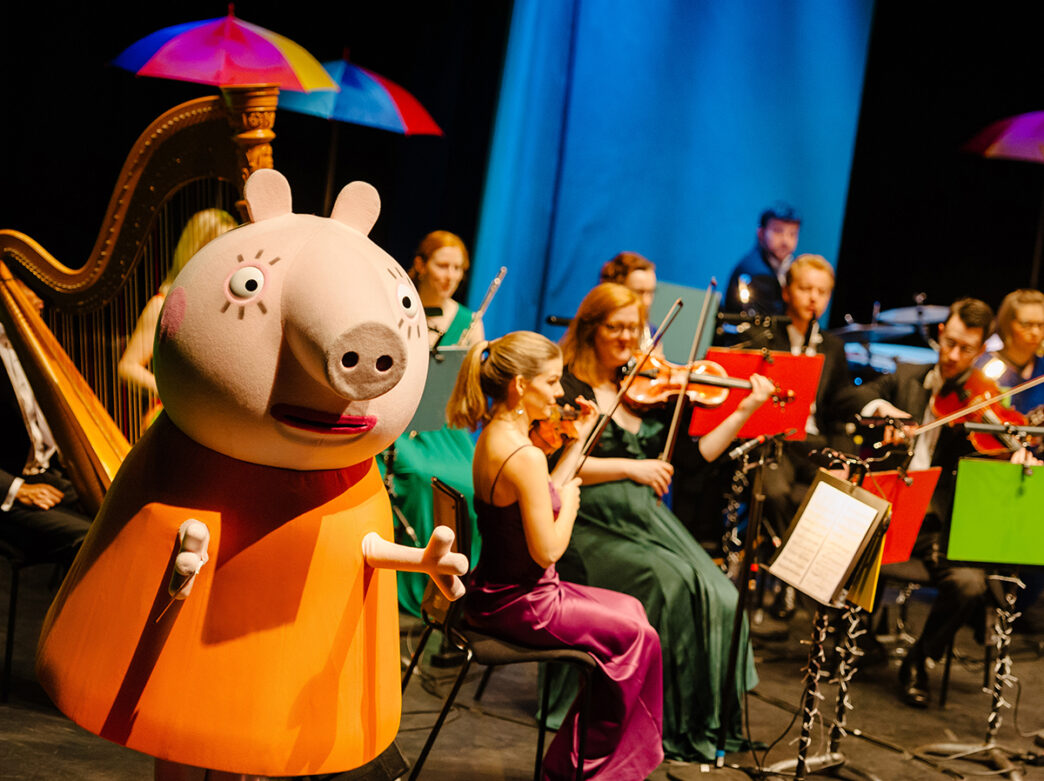 A large Peppa Pig character stands onstage in front of a seated orchestra, with musicians playing strings and colorful umbrellas hang above the performance.