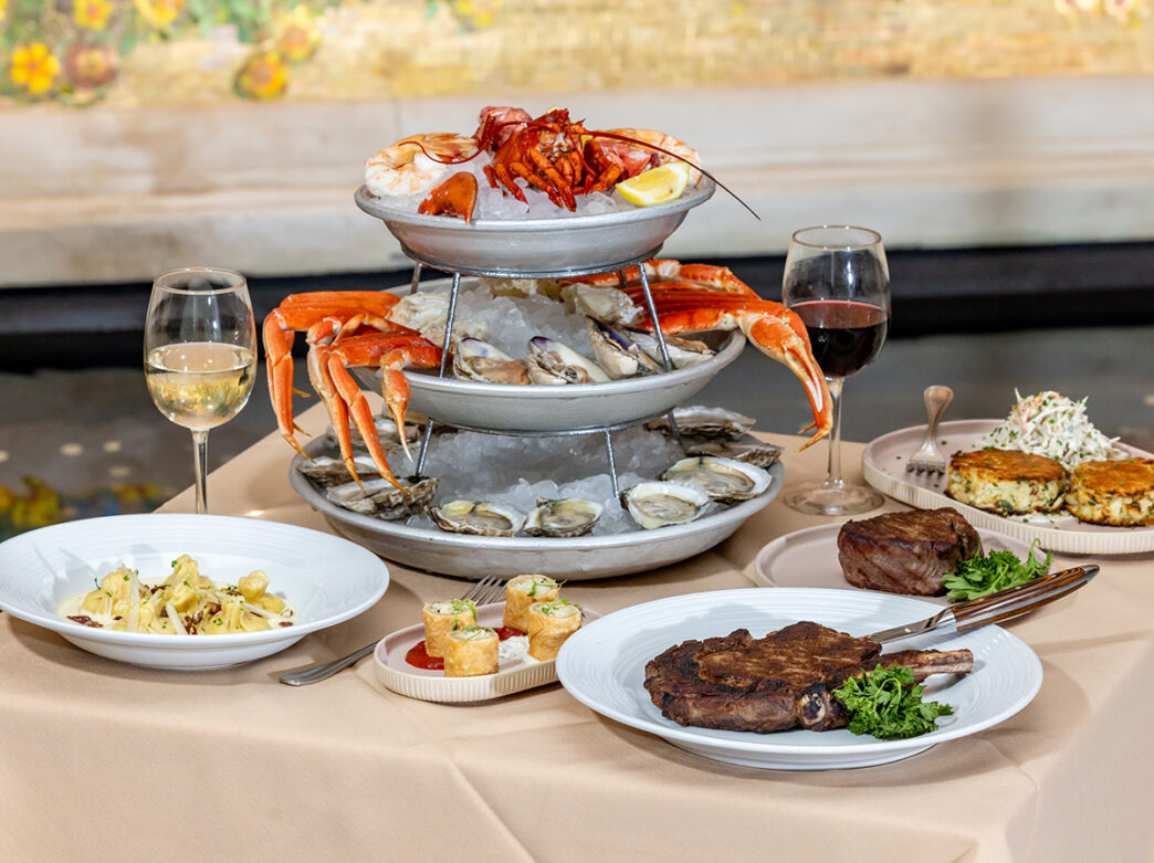 A seafood tower filled with oysters, shrimp and lobster sits on a table alongside wine glasses, steak, pasta and other dishes in a restaurant setting.