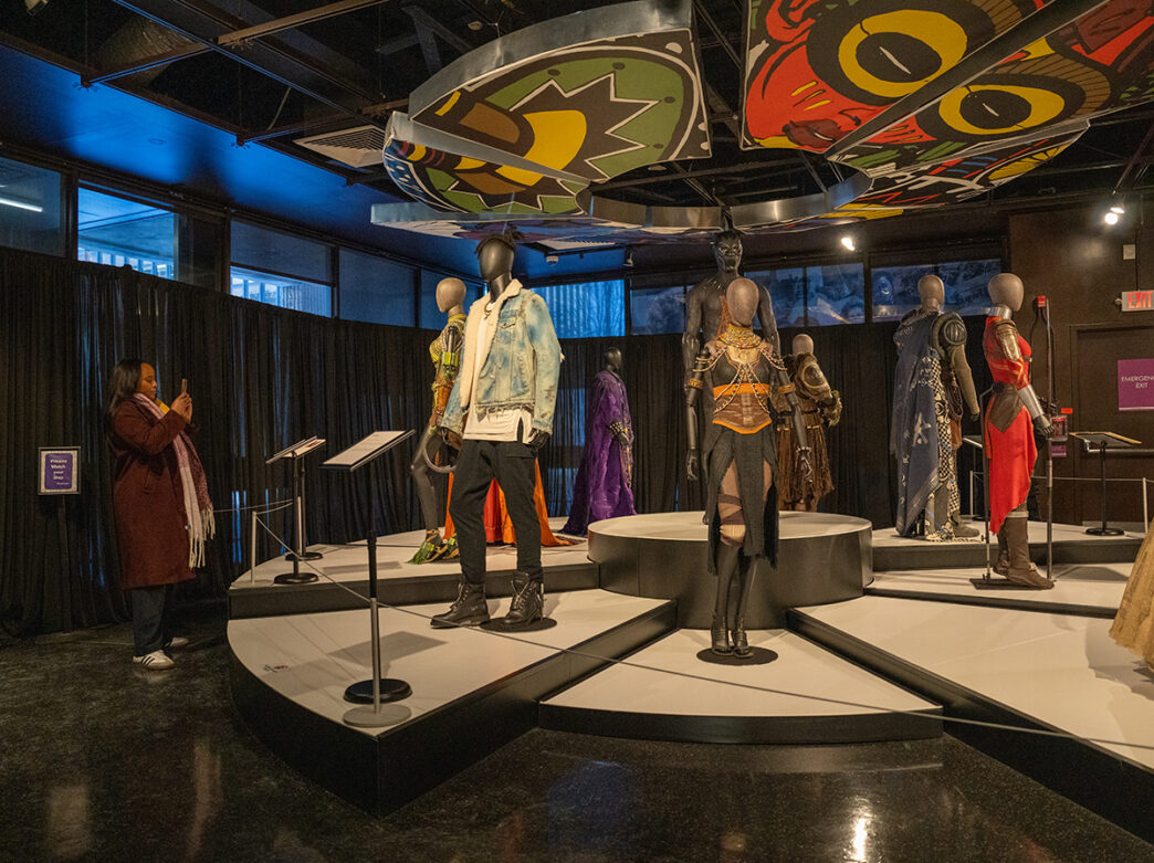Mannequins dressed in detailed Afrofuturist costumes stand on a circular platform inside a dimly lit gallery, as a visitor takes a photograph of the exhibit.