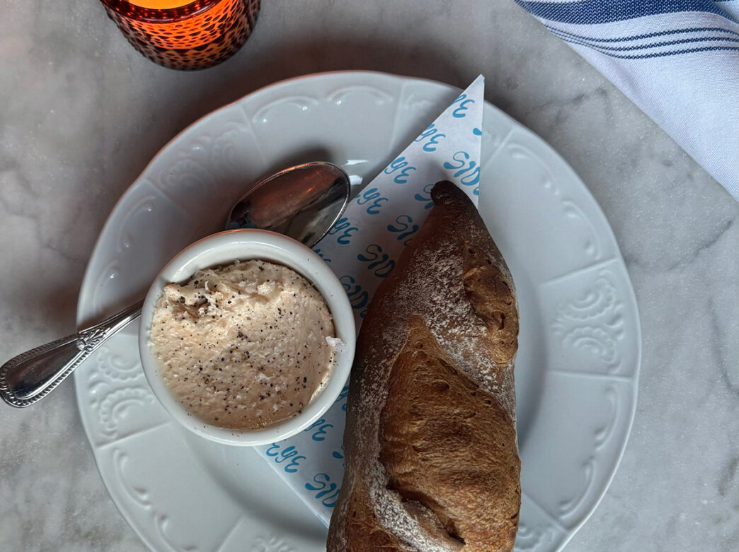A white plate holds a crusty loaf of bread alongside a small ramekin of whipped butter sprinkled with black pepper, set on a marble tabletop.