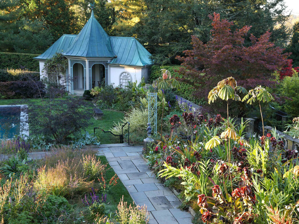 A lush garden scene at Chanticleer with vibrant green and burgundy folliage and a charming green-roofed cottage.