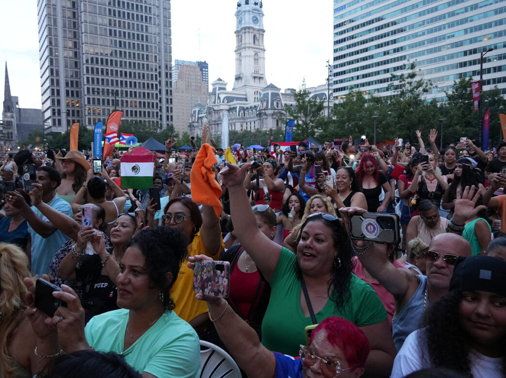 Une foule nombreuse et diversifiée applaudit et enregistre une performance en plein air devant le City Hall de Philadelphie, avec des drapeaux, des mains levées et des téléphones visibles dans tout le public.