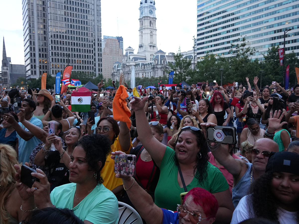 A large, diverse crowd cheers and records a live outdoor performance in front of Philadelphia's City Hall, with flags, raised hands and phones visible throughout the audience.