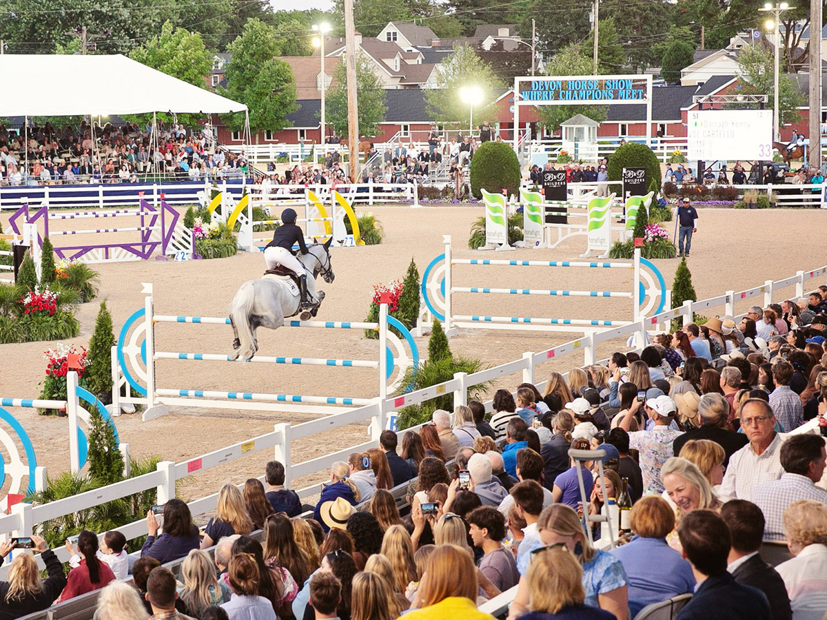 A light gray horse and its sharply dressed rider jump above an obstacle at the Devon Horse Show while the packed crowd in the grandstands watches.
