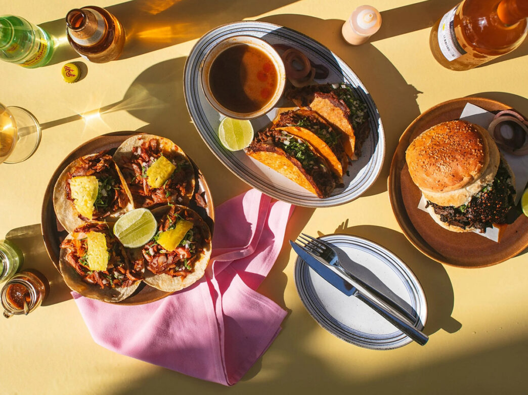 An overhead image of a spread of dishes sitting on a pale yello table. The spread includes a sandwich piled high with grilled meat, a plate of three birria tacos and a plate with four tacos al pastor. Bottles of beer and Topo Chico can also be seen on the table.