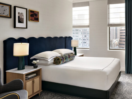Chambre moderne du Kimpton Hotel Monaco Philadelphia avec un lit en peluche doté d'une tête de lit capitonnée de couleur marine, des lampes de chevet, une chaise grise et deux fenêtres donnant sur la ville.