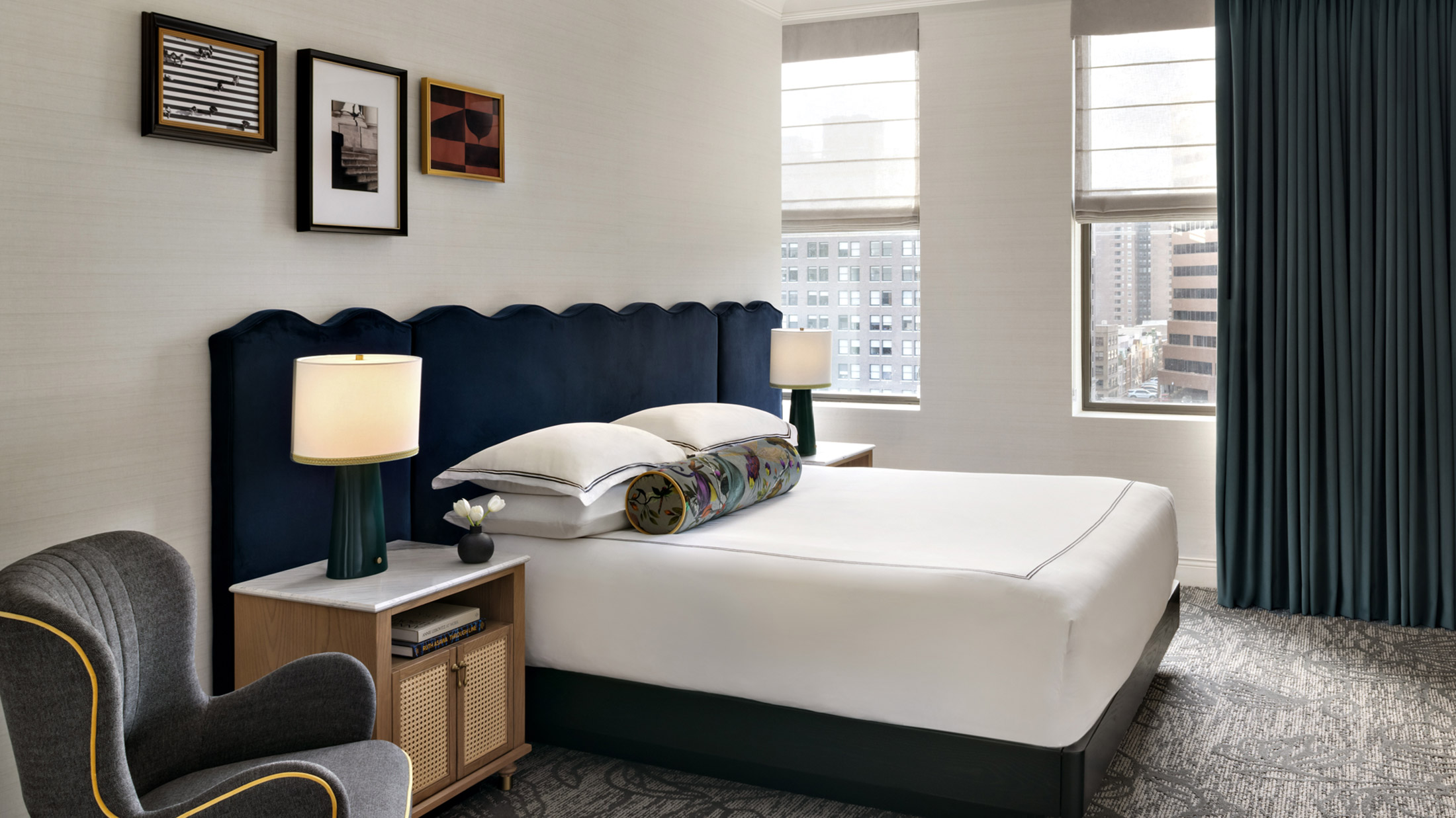 Moderna habitación del Kimpton Hotel Monaco Philadelphia con una lujosa cama con cabecera tapizada en azul marino, lámparas de noche, una silla gris y dos ventanas con vistas a la ciudad.