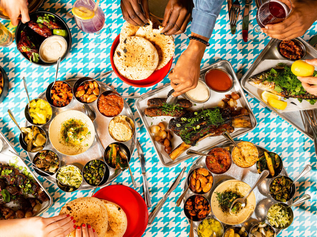A spread of Laser Wolf menu items set against a teal and white houndstooth pattern. The dishes include fries with ketchup, grilled meats, whole fish with herbs and lemon, pita, an assortment of hummus and other dips. Diners' hands can be seen reaching for food.
