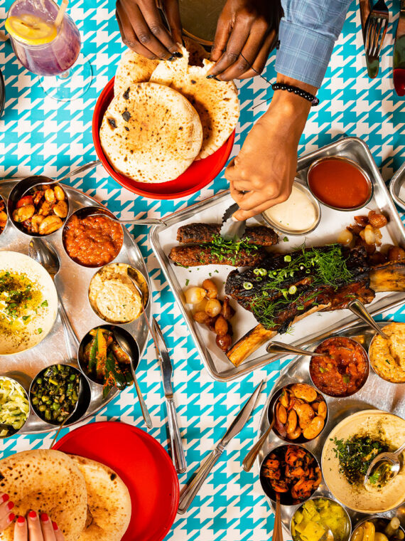 A spread of Laser Wolf menu items set against a teal and white houndstooth pattern. The dishes include fries with ketchup, grilled meats, whole fish with herbs and lemon, pita, an assortment of hummus and other dips. Diners' hands can be seen reaching for food.
