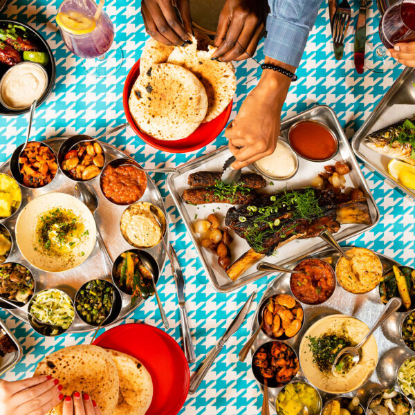 A spread of Laser Wolf menu items set against a teal and white houndstooth pattern. The dishes include fries with ketchup, grilled meats, whole fish with herbs and lemon, pita, an assortment of hummus and other dips. Diners' hands can be seen reaching for food.