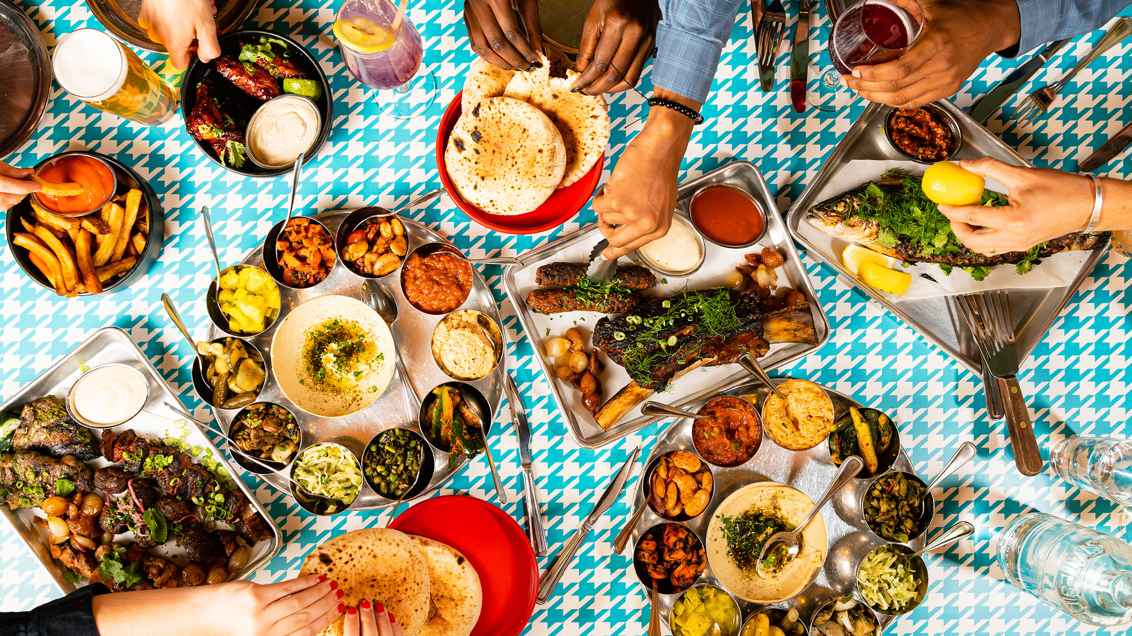 Una variedad de platos del menú de Laser Wolf sobre un fondo con un estampado de pata de gallo en verde azulado y blanco. Los platos incluyen papas fritas con ketchup, carnes a la parrilla, pescado entero con hierbas y limón, pan de pita, una variedad de hummus y otras salsas. Se puede ver a los comensales con las manos extendidas para tomar la comida.