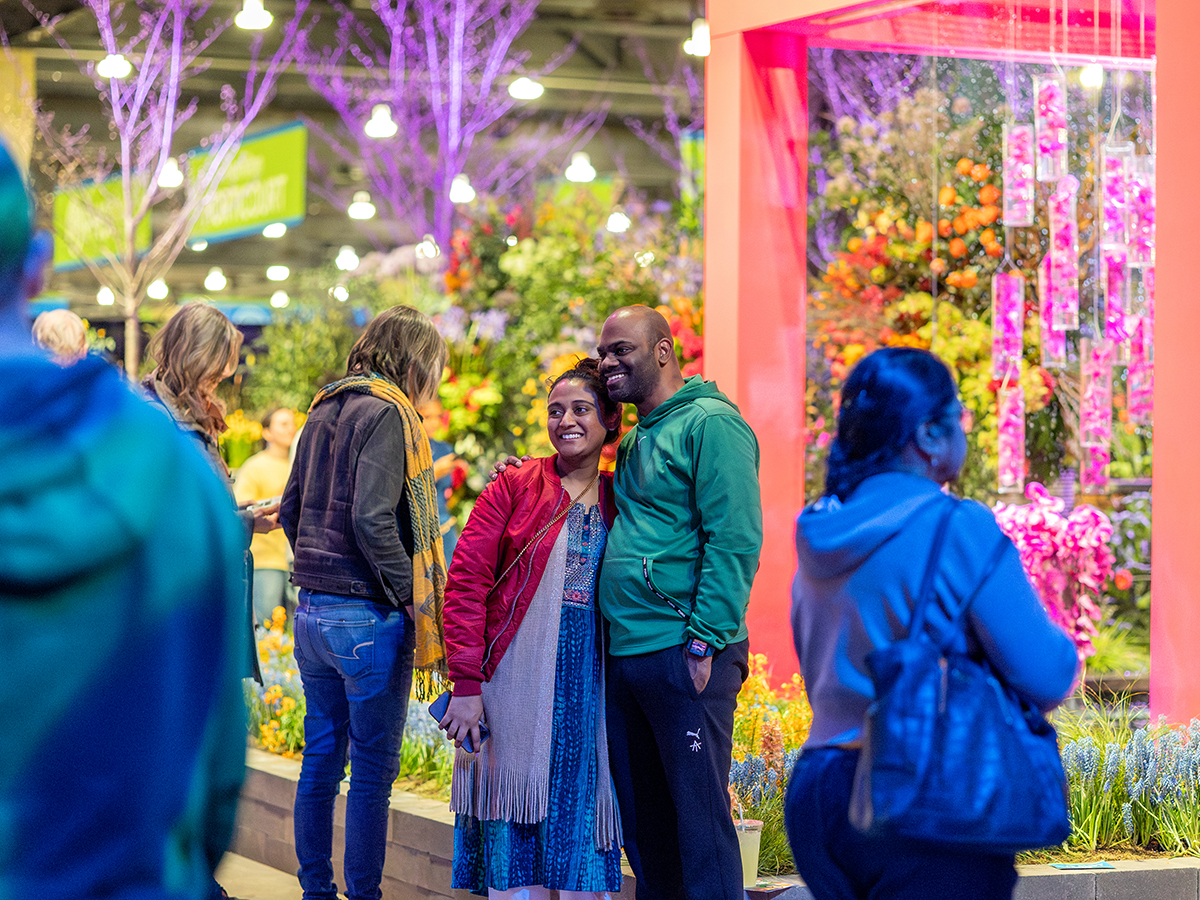 A couple poses for a photo at the PHS Philadelphia Flower Show, surrounded by colorful floral displays and glowing pink and purple lights.