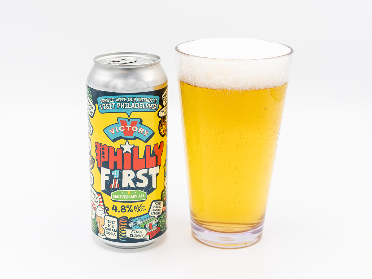 A can of "Philly First" Anniversary Ale stands besides a full pint glass of golden beer with a foamy head against a white background.