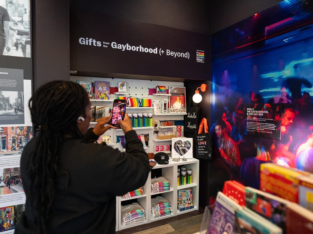 A visitor takes a photo inside the Philly Pride Visitor Center, standing beside shelves of rainbow-themed merchandise under a sign reading “Gifts from the Gayborhood (+ Beyond).”
