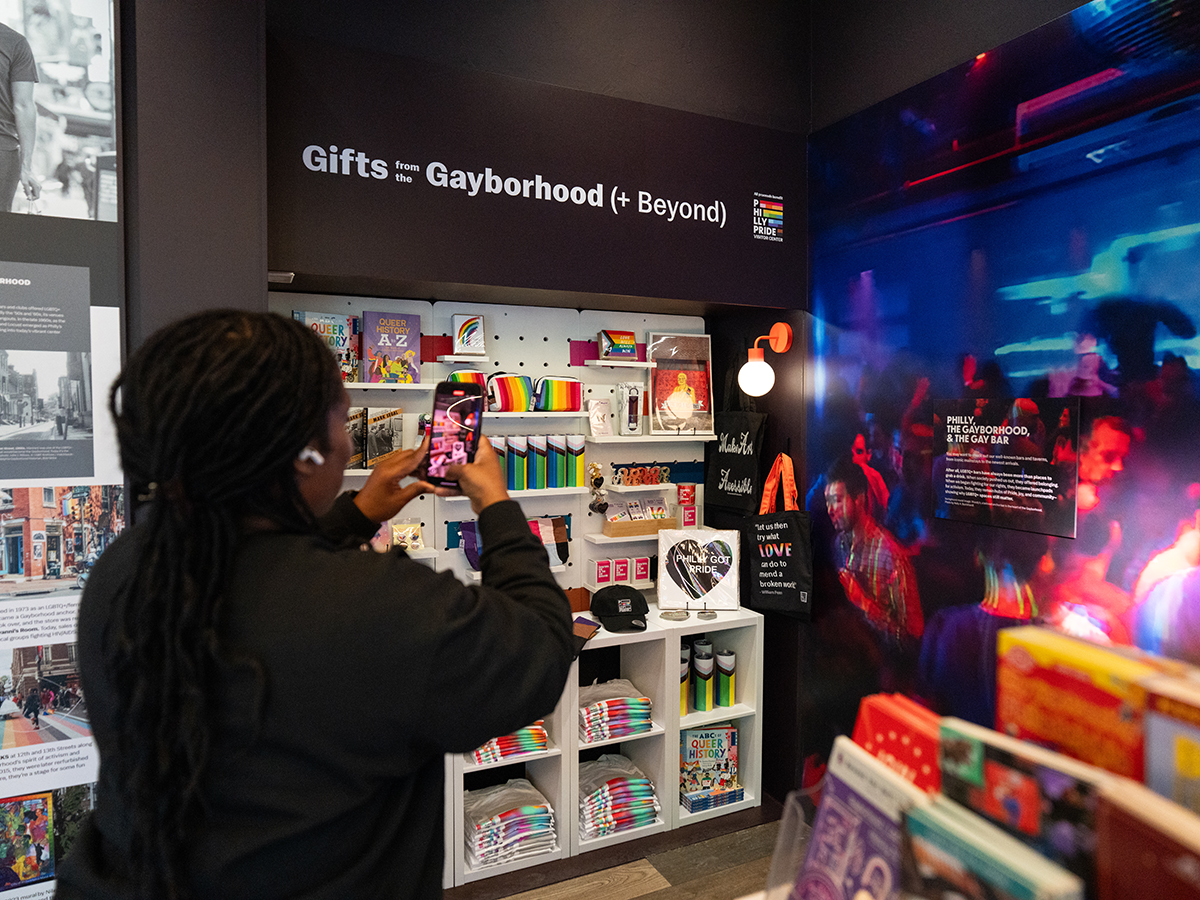 A visitor takes a photo inside the Philly Pride Visitor Center, standing beside shelves of rainbow-themed merchandise under a sign reading “Gifts from the Gayborhood (+ Beyond).”