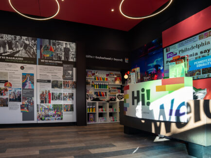 An interior view of the Philly Pride Visitor Center showing colorful displays on LGBTQ+ history, a bright welcome desk and Pride merchandise displays beneath modern circular light fixtures.
