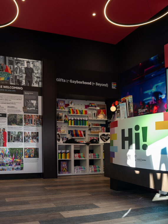 An interior view of the Philly Pride Visitor Center showing colorful displays on LGBTQ+ history, a bright welcome desk and Pride merchandise displays beneath modern circular light fixtures.