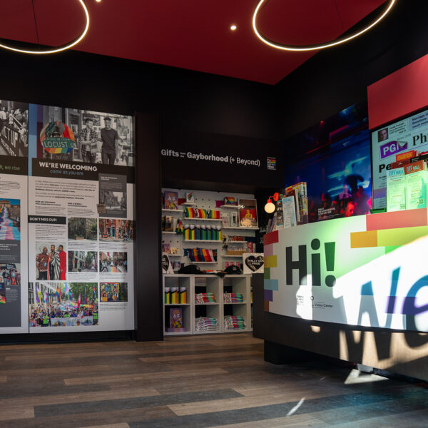 Uma vista do interior do Pride Visitor Center do Philly mostrando displays coloridos sobre a história do LGBTQ+, um balcão de boas-vindas bem iluminado e displays de mercadorias do Pride sob modernas luminárias circulares.