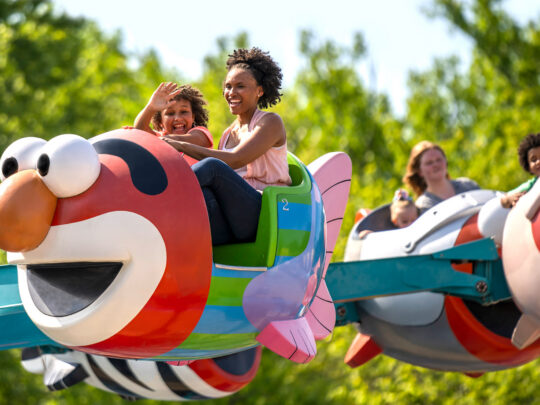 A mother and daughter smile and wave from inside a colorful, Elmo-shaped ride at Sesame Place.