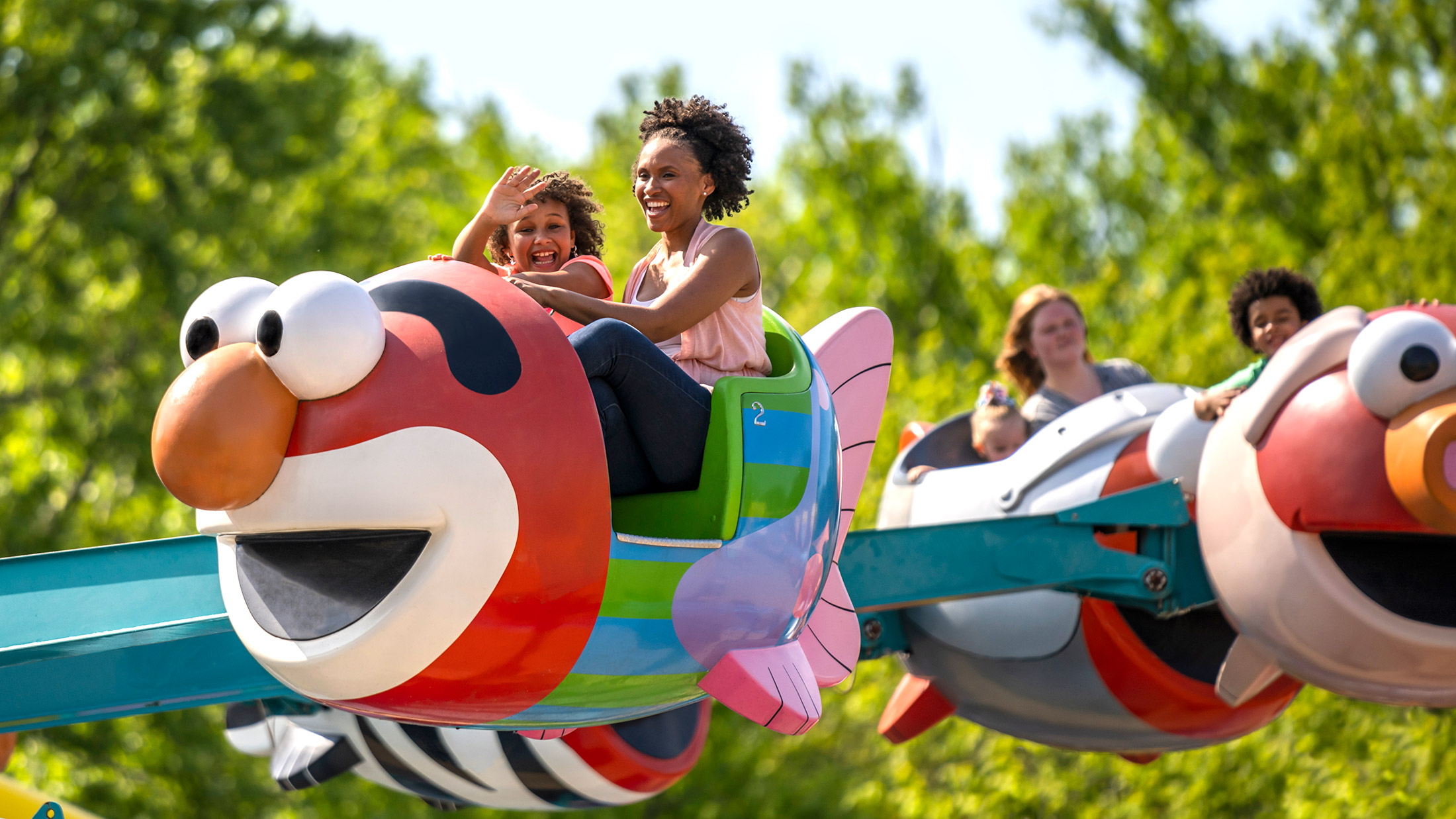 Een moeder en dochter lachen en zwaaien vanuit een kleurrijke attractie in de vorm van Elmo op Sesame Place.