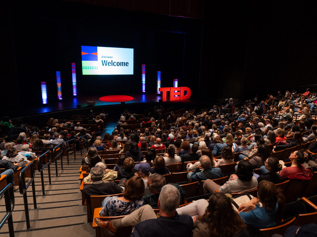 A packed theater audience sits facing a brightly lit stage, where a screen reads "Welcome" alongside TED Democracy branding and colorful pillars.
