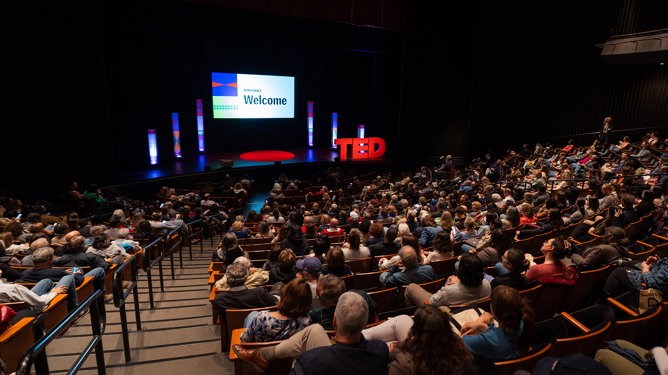A packed theater audience sits facing a brightly lit stage, where a screen reads "Welcome" alongside TED Democracy branding and colorful pillars.