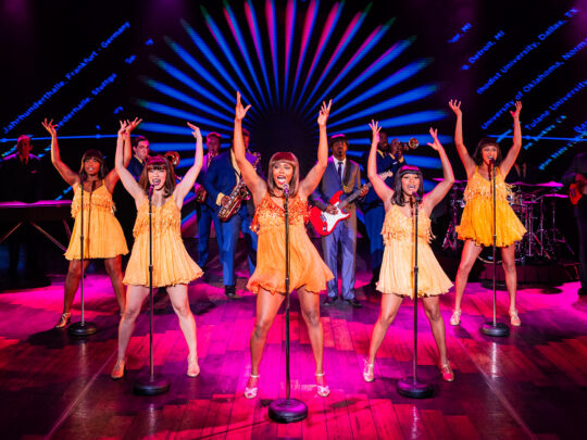 Women dressed in yellow dance on a pink stage for a performance of the Tina Turner musical