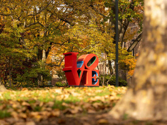 Una réplica de la emblemática estatua roja LOVE de la Universidad de Pensilvania rodeada de árboles otoñales en tonos naranjas y verdes.