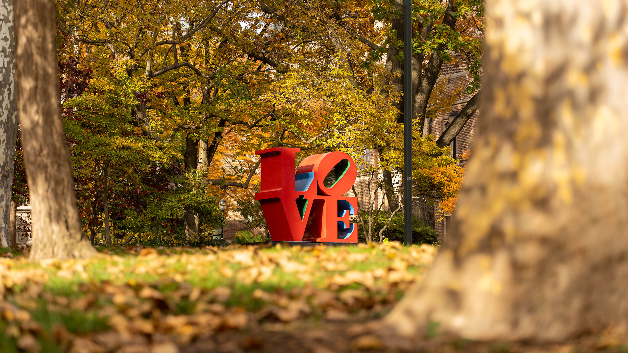 A replica statue of the iconic red LOVE statue at the University of Pennsylvania is surrounded by autumnal trees in shades of orange and green.