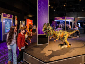 Three children lean forward excitedly as a small green animatronic dinosaur snarls on a raised platform inside the Universal Theme Parks exhibit at The Franklin Institute.