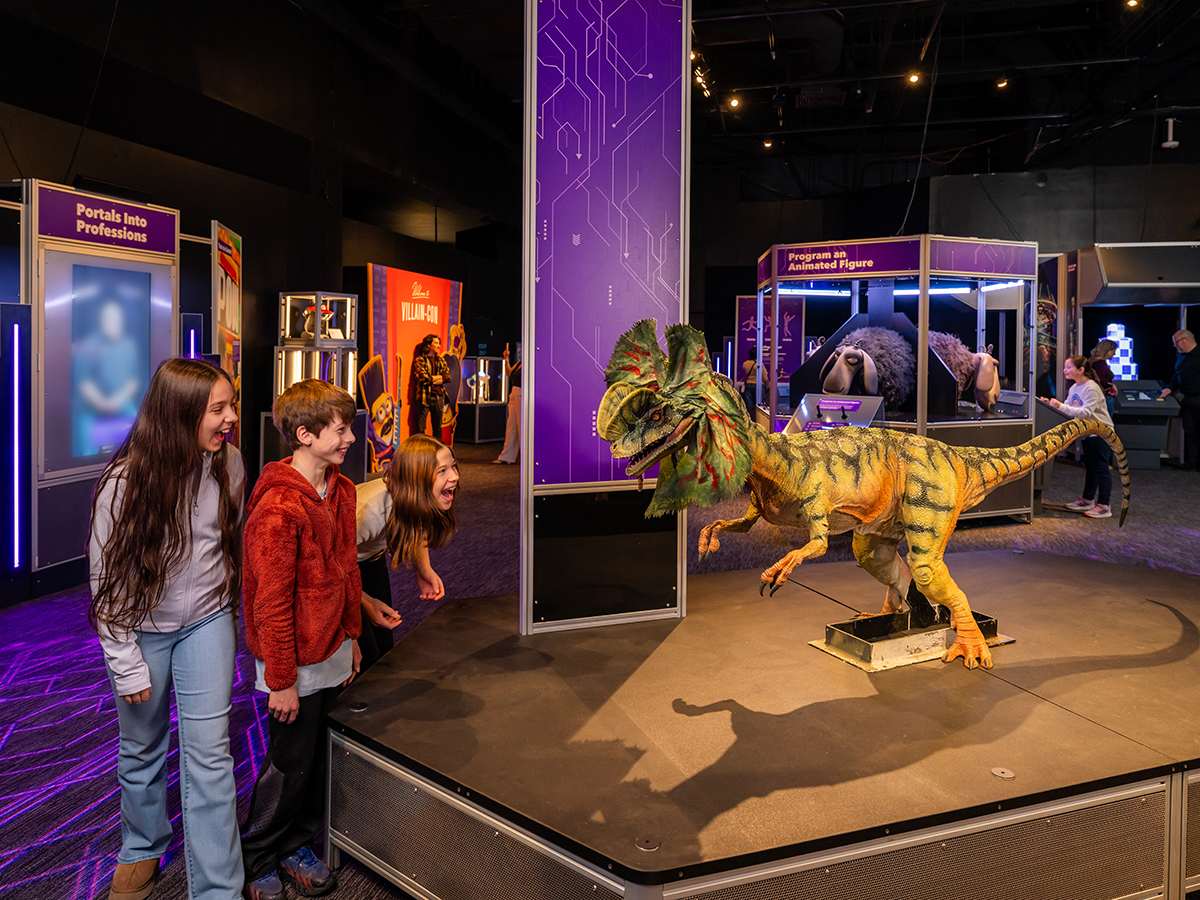 Three children lean forward excitedly as a small green animatronic dinosaur snarls on a raised platform inside the Universal Theme Parks exhibit at The Franklin Institute.