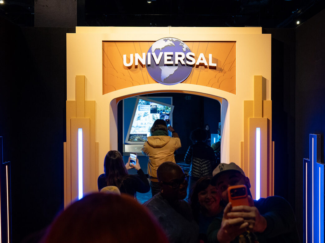 Os visitantes tiram fotos sob um arco de entrada brilhante da "Universal" dentro da exposição Universal Theme Parks no The Franklin Institute.