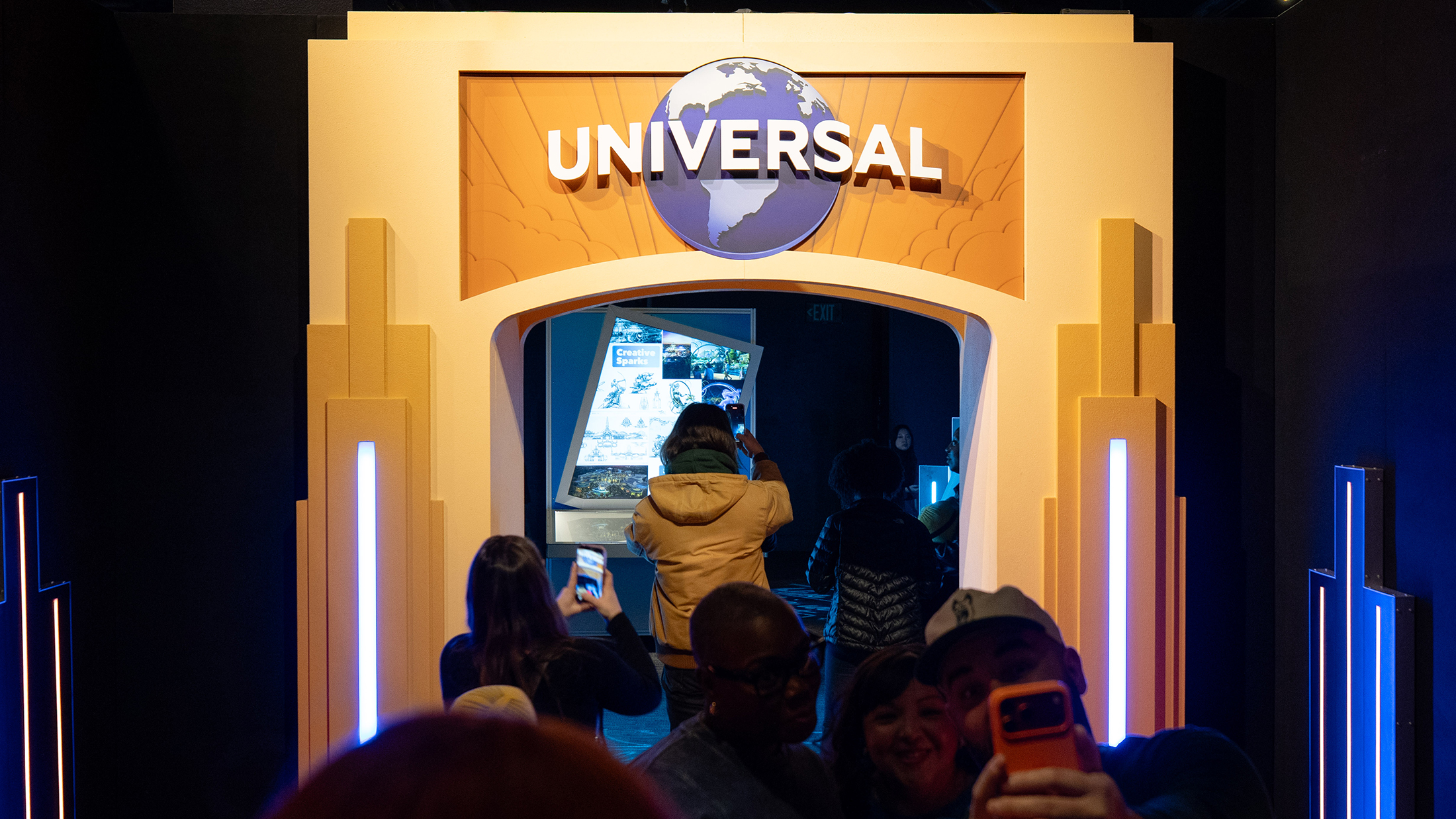Los visitantes se toman fotos bajo un arco luminoso con la palabra "Universal" en la entrada de la exposición Universal Theme Parks en The Franklin Institute.