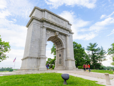 Los visitantes pasean bajo un imponente arco de piedra que se alza en medio de un césped verde y árboles frondosos en Valley Forge.