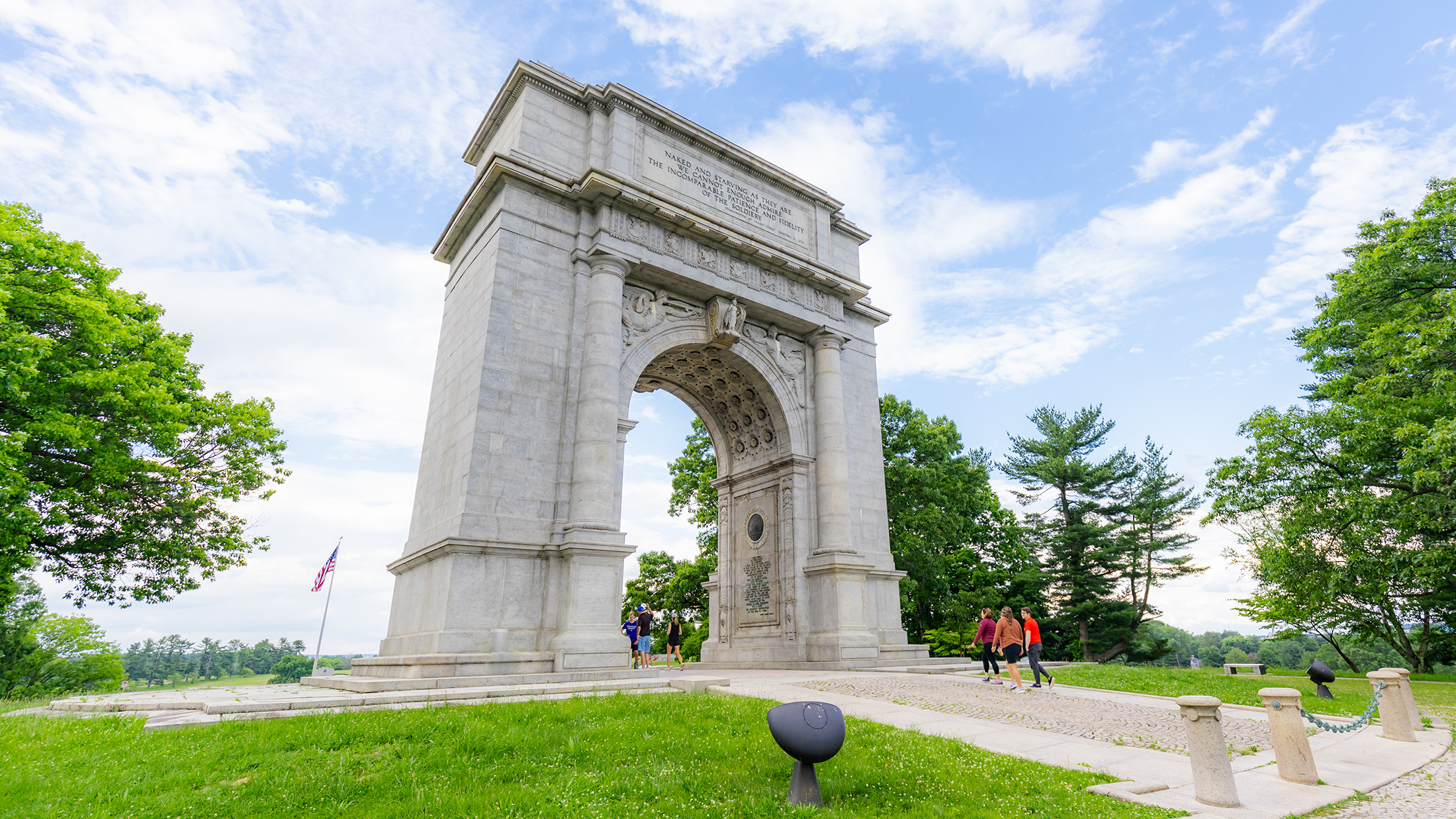 Os visitantes passeiam sob um imponente arco de pedra, situado em meio a um gramado verdejante e árvores frondosas em Valley Forge.