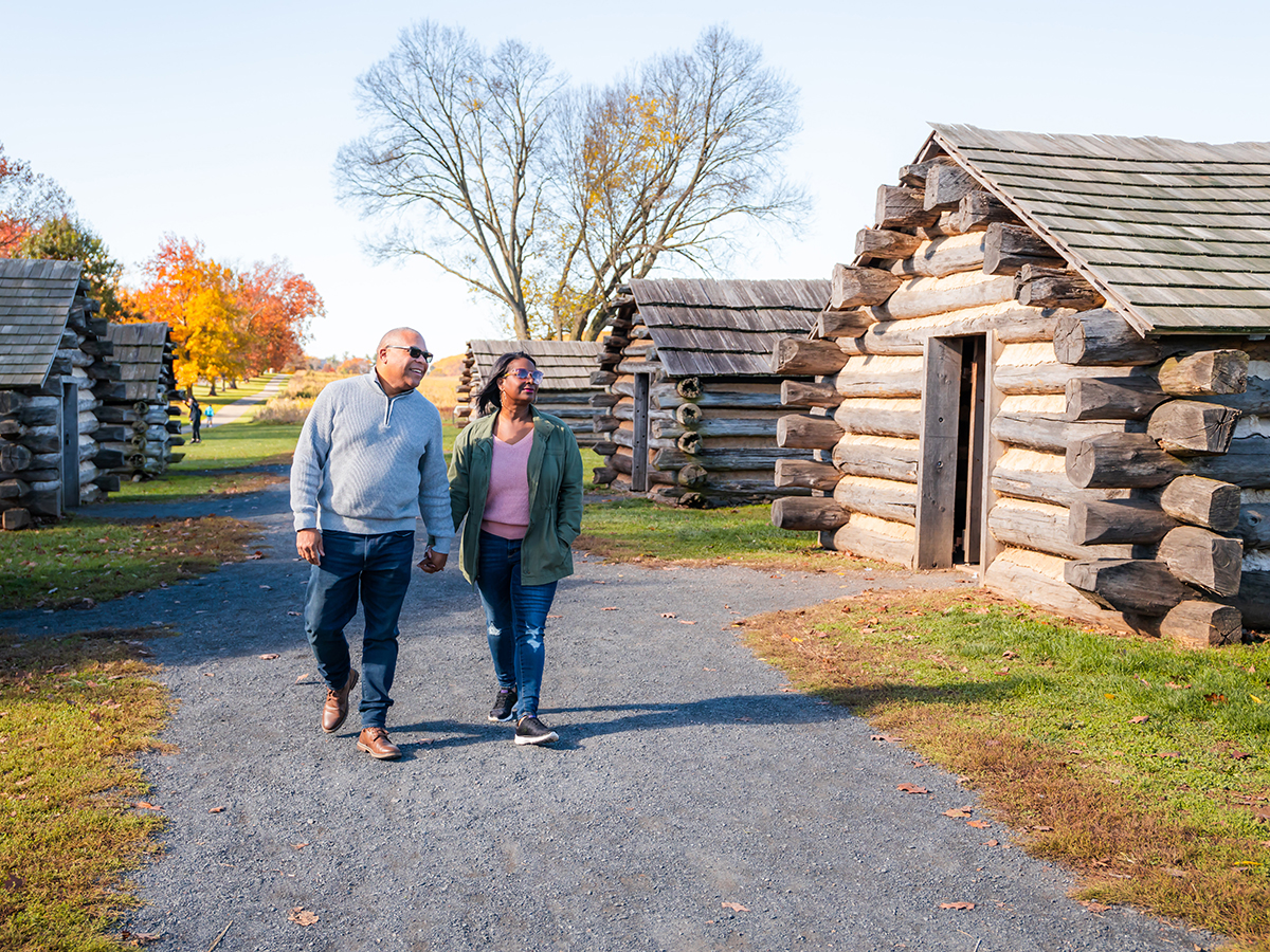 A couple walks hand in hand past restored Revolutionary War log cabins at Valley Forge, surrounded by colorful fall foliage under a clear sky.
