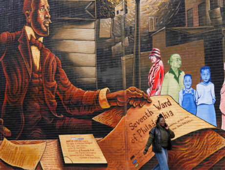 A pedestrian walking by a large mural on the side of a building, featuring a portrait of WEB Du Bois, historic documents and colorful community members.