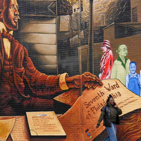 Un piéton passe devant une grande peinture murale sur le côté d'un bâtiment, représentant un portrait de WEB Du Bois, des documents historiques et des membres de la communauté hauts en couleur.