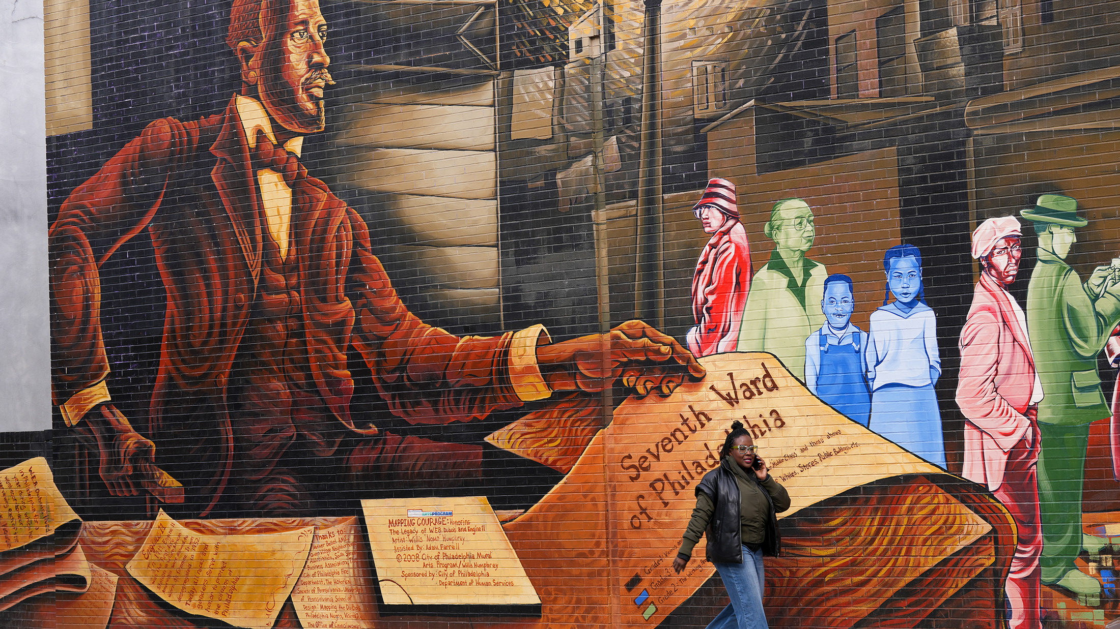 Un piéton passe devant une grande peinture murale sur le côté d'un bâtiment, représentant un portrait de WEB Du Bois, des documents historiques et des membres de la communauté hauts en couleur.