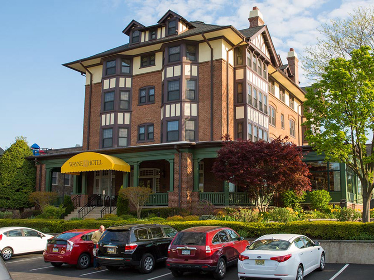 Cars parked outside the brick facade of the Wayne Hotel. A bright yellow awning bearing its name and green patio can be seen.