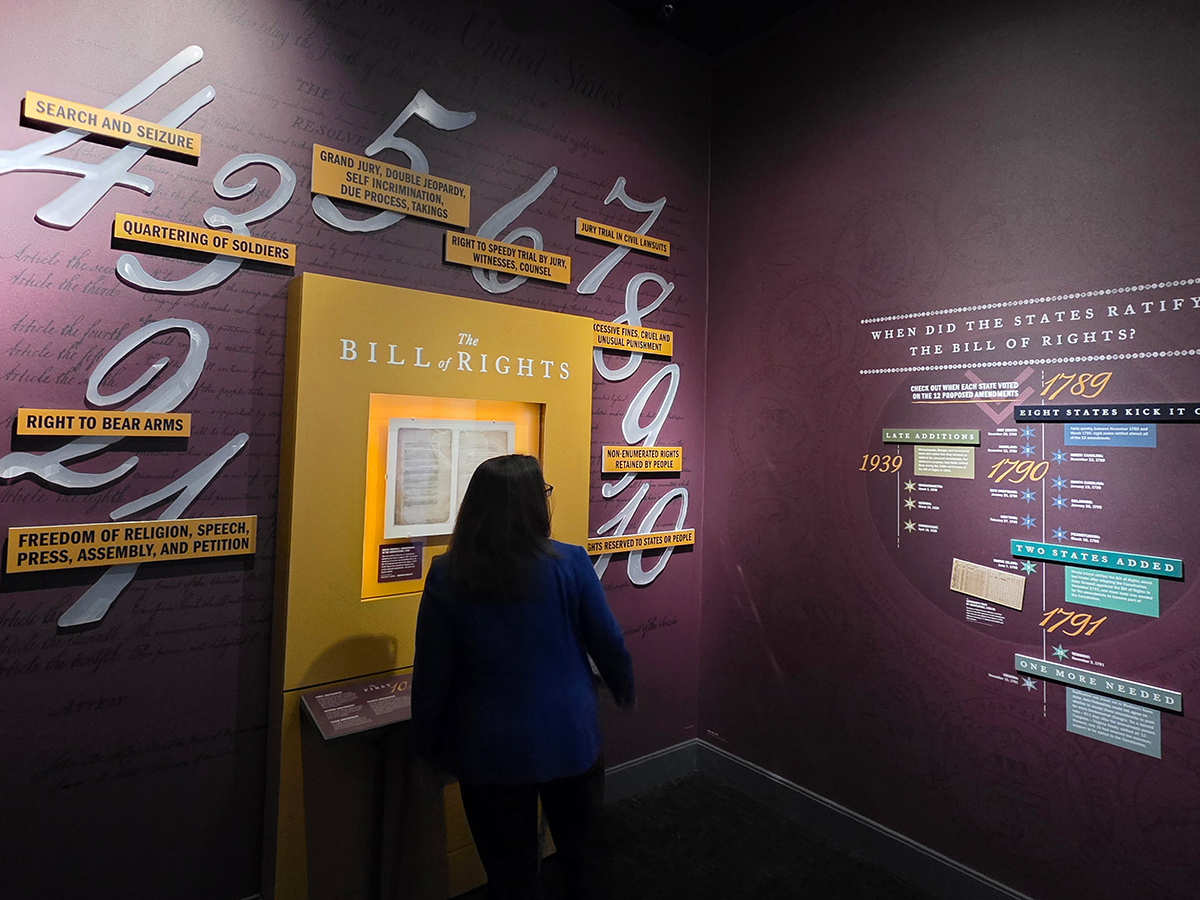 A visitor reads the Bill of Rights displayed in a bright yellow case in a museum gallery, surrounded by large wall graphics listing freedoms such as speech, religion and right to bear arms.
