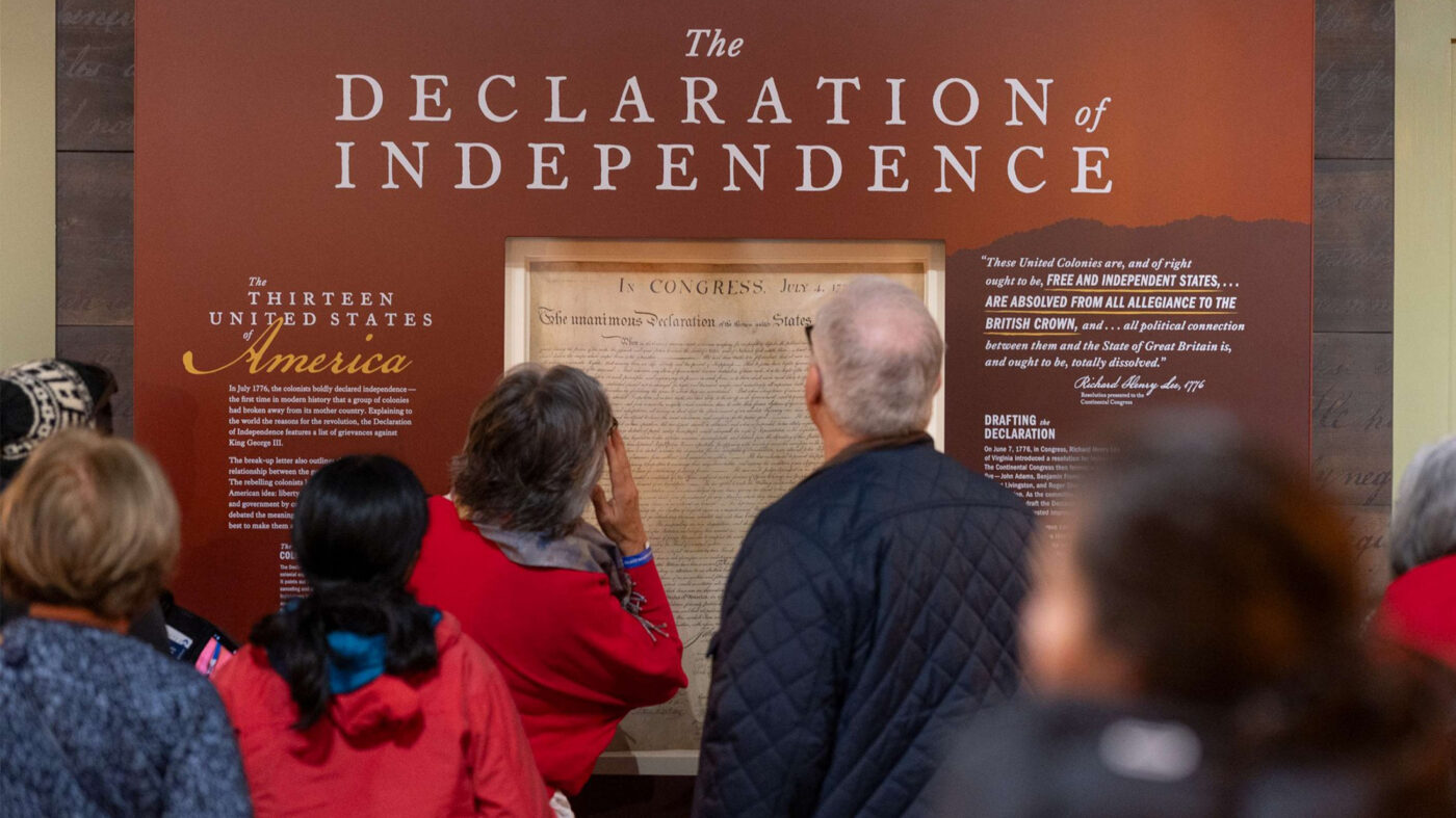 Visitors stand in front of a large wall display featuring the Declaration of Independence, reading text panels and viewing a frame reproduction of the historic document in a museum gallery.