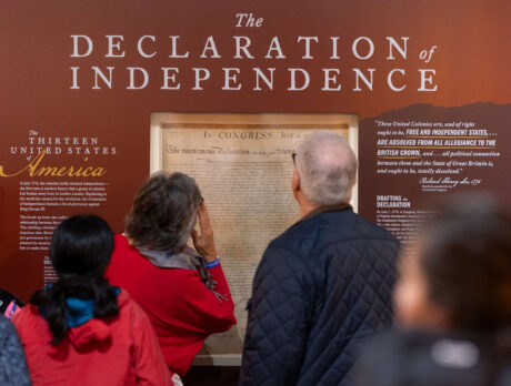 Visitors stand in front of a large wall display featuring the Declaration of Independence, reading text panels and viewing a frame reproduction of the historic document in a museum gallery.
