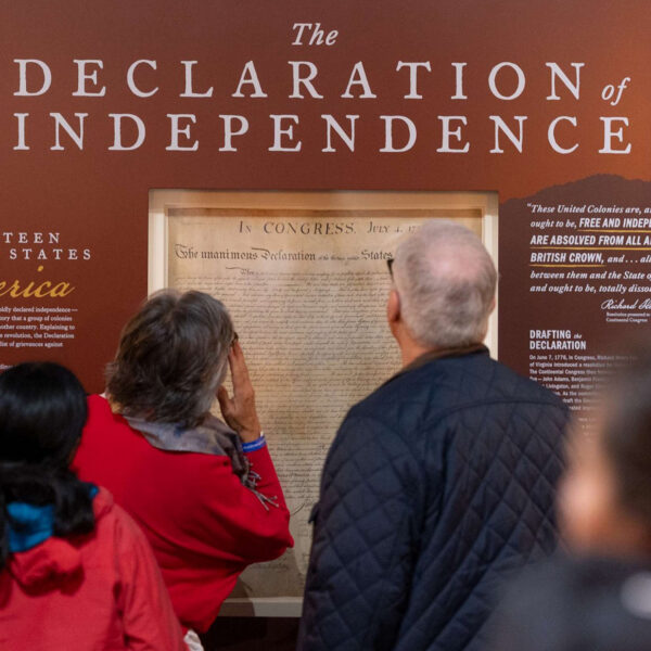 Visitors stand in front of a large wall display featuring the Declaration of Independence, reading text panels and viewing a frame reproduction of the historic document in a museum gallery.