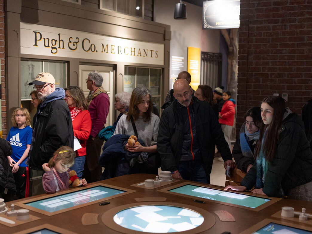 Families gather around a large interactive touchscreen table inside a recreated storefront labeled "Pugh & Co. Merchants," exploring hands-on elements together.