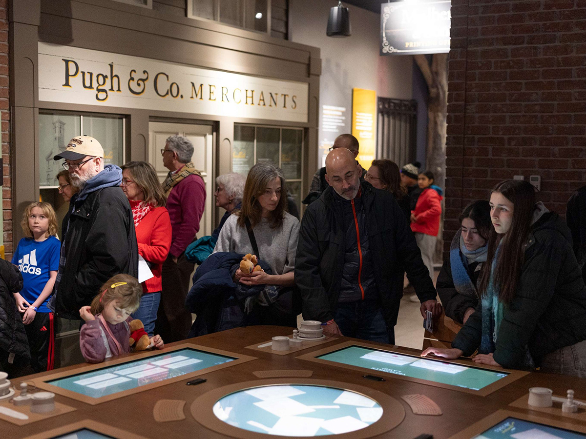 Families gather around a large interactive touchscreen table inside a recreated storefront labeled "Pugh & Co. Merchants," exploring hands-on elements together.