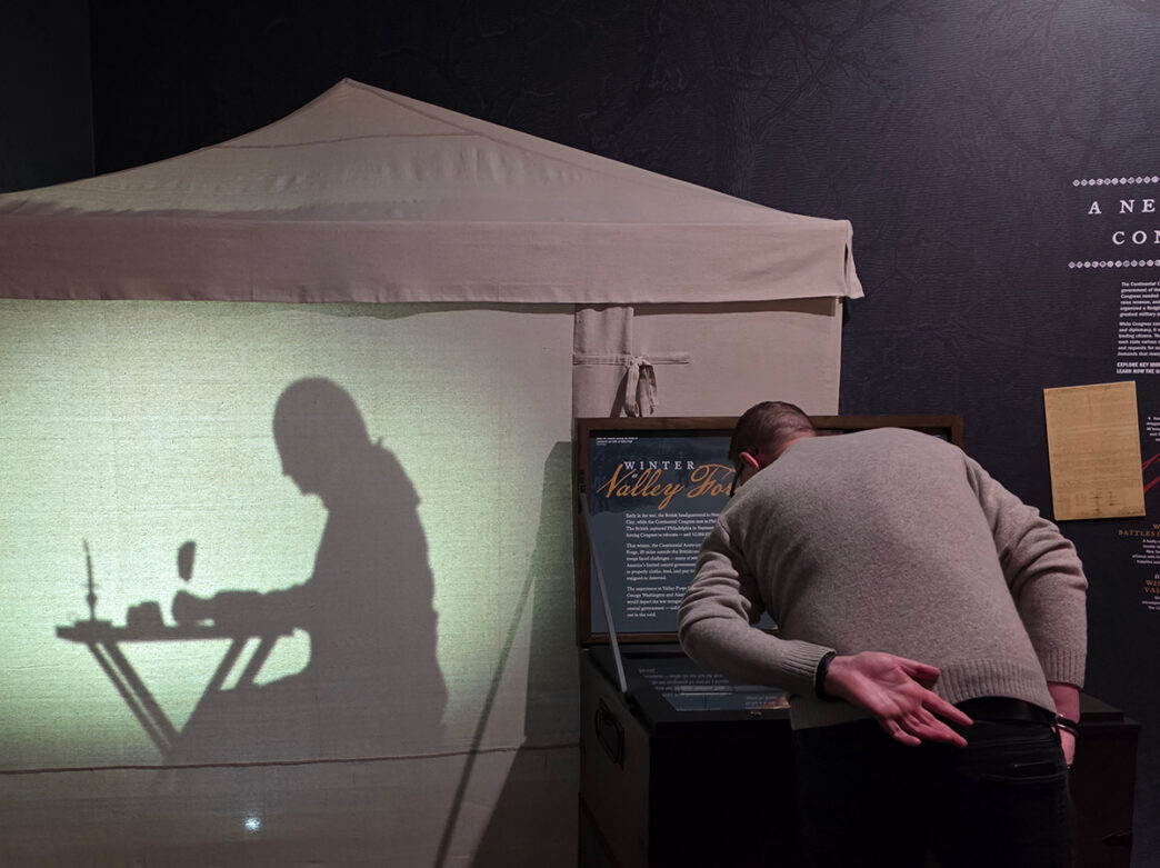 A museum visitor leans over an interactive display while, on the wall beside him, a shadow silhouette shows a revolutionary war soldier writing at a small camp desk inside a tent.