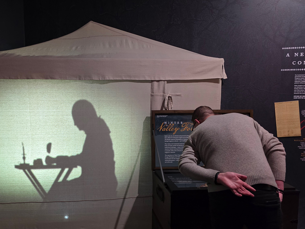 A museum visitor leans over an interactive display while, on the wall beside him, a shadow silhouette shows a revolutionary war soldier writing at a small camp desk inside a tent.