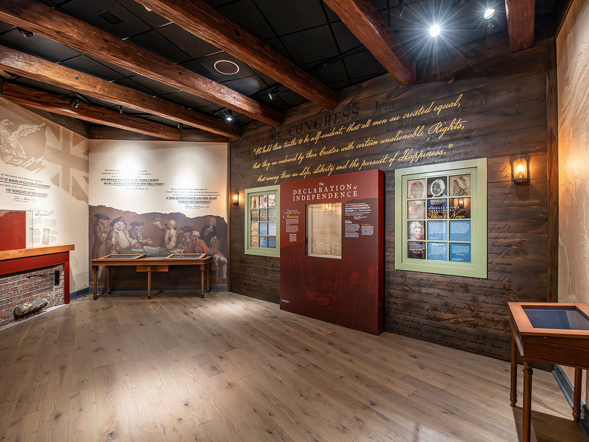 A wide angle view of a wood-paneled gallery displaying the Declaration of Independence at the center, surrounded by interpretive panels, historic imagery and warm lighting beneath exposed wooden beams.